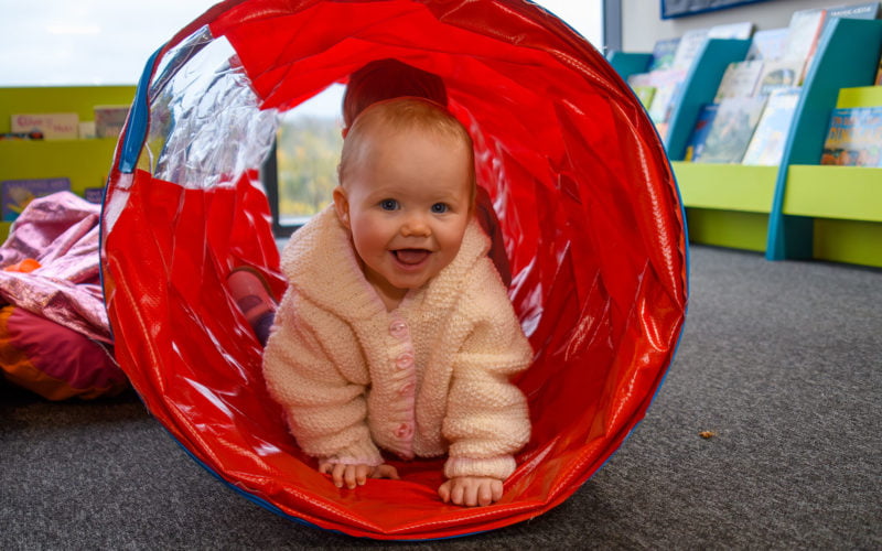 A smiling white baby wearing a pink jumper crawls through a res play tunnel looking at the camera