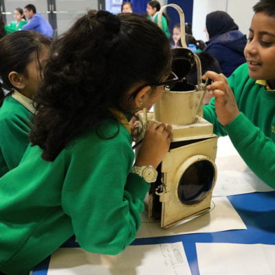 School children dressed in green uniform inspect a cream coloured Victorian railway lamp