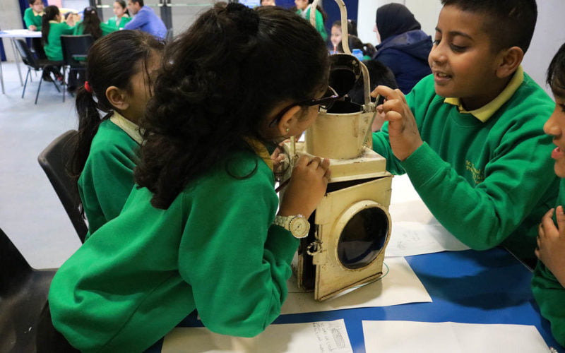 School children dressed in green uniform inspect a cream coloured Victorian railway lamp