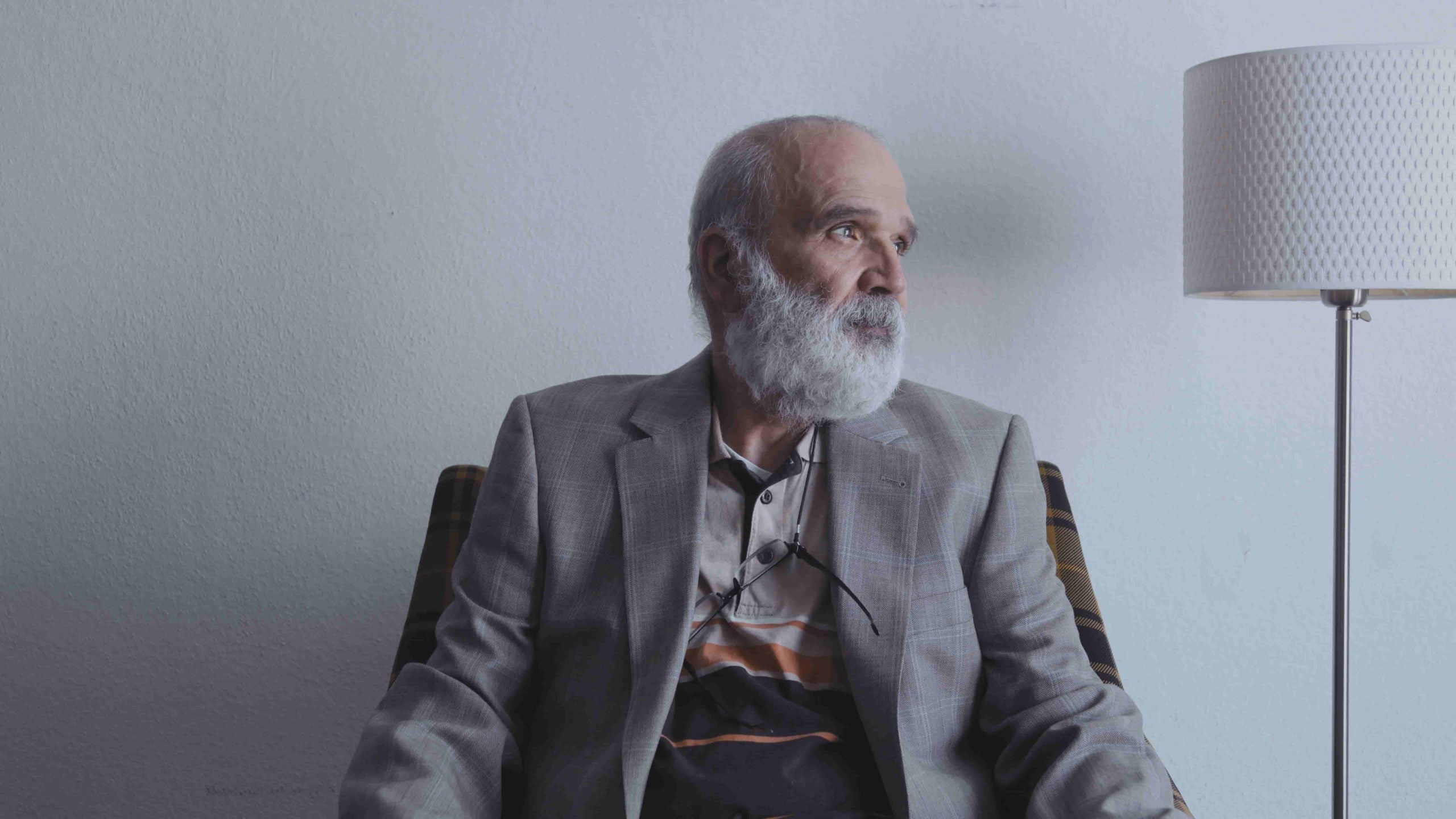 A bearded in a grey suite man sits looking to the side in front of a blank white wall with a white lamp next to him.