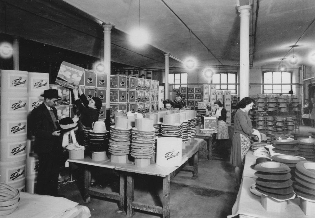 Photograph showing an interior view at Failsworth Hats, including women packing hats into boxes and a man checking the quality of one of the hats.
Image reference: P21006