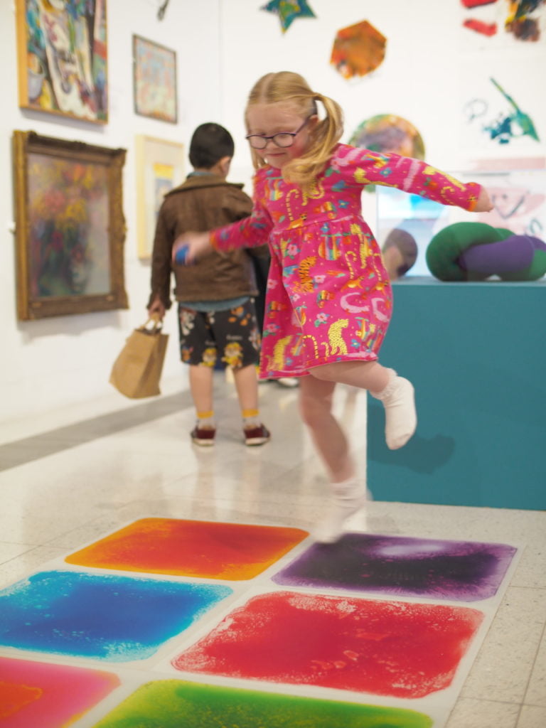 A little girl wearing a pink dress and glasses jumping on some coloured squares