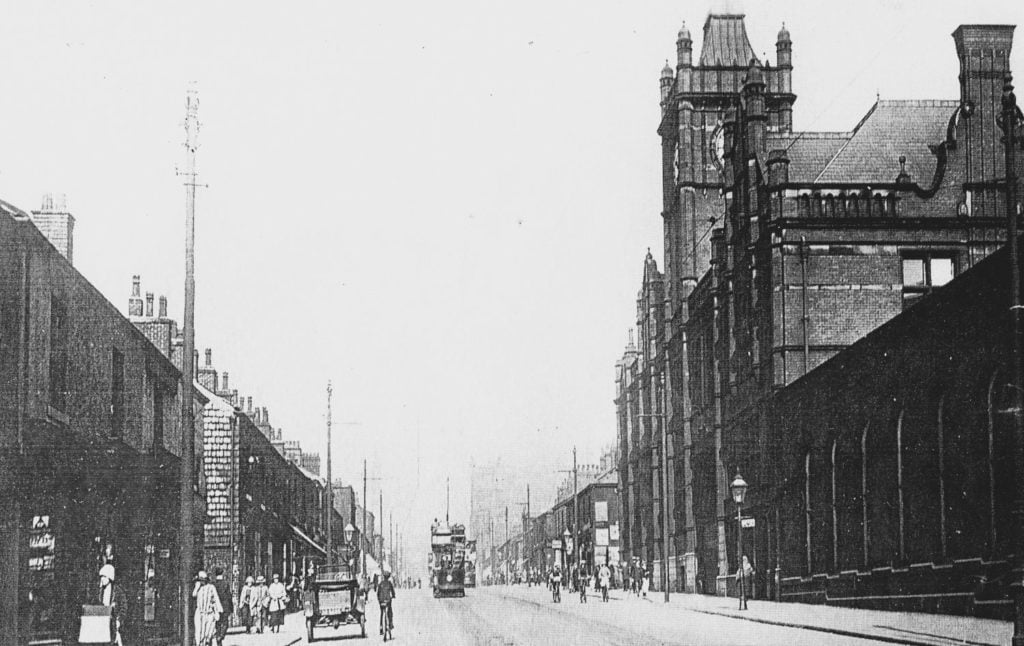 Photograph of Asa Lees Mill with it's famous clocktower from 1906. Shown is the road in front with pedestrians, a tram, cyclists and a motorcar.