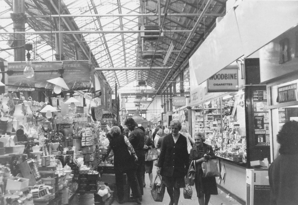 Photograph of the inside of Victoria Market Hall showing shoppers, a newsagents and other stalls.