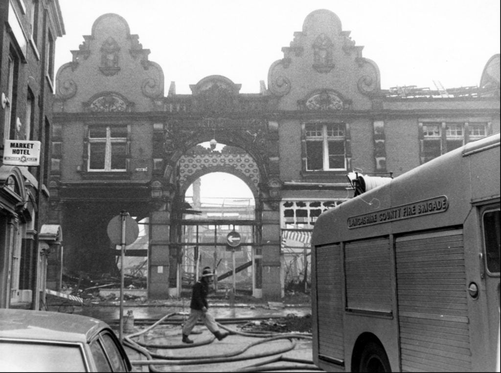 Photograph of Lancashire County Fire Brigade fire truck in foreground and façade of the burnt down Victoria Market Hall in the the background with firefighter walking across the street.