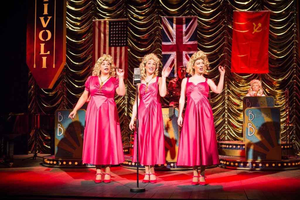 Three women in pink dresses with blonde hair singing with America, Great Britain and Russian flag behind.