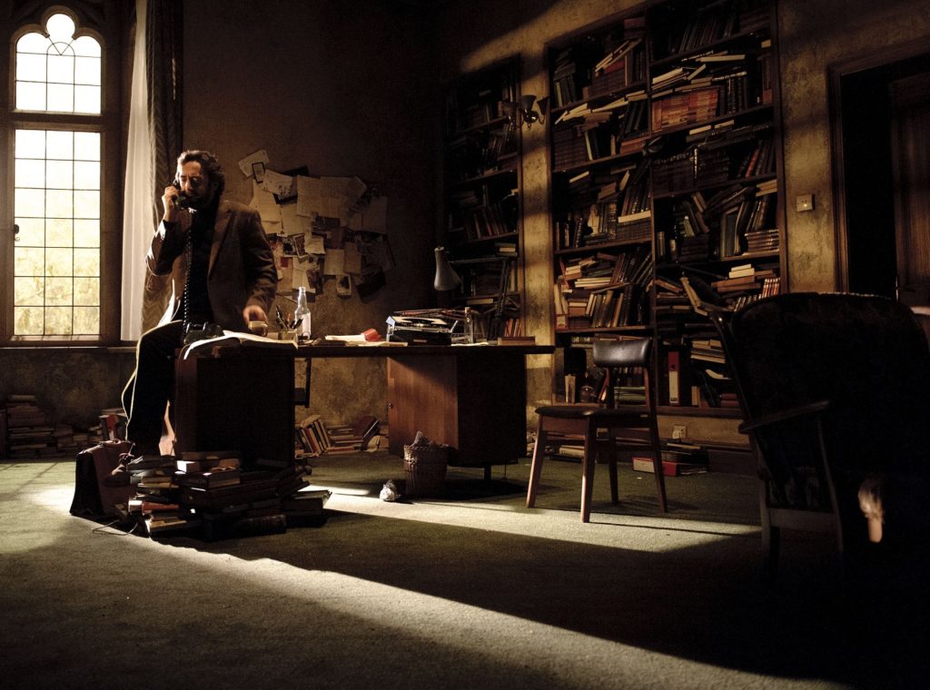 Set showing actor sat on desk using the phone surrounded by bookshelves.