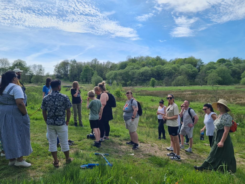 Photograph of a group of people at the Northern Roots site on a sunny day. The group is standing in a field listening to someone speak to them.