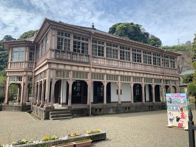 Photo of a building built in a mixture of European and Japanese style. It is two stories high, with a balcony running around the upper floor. It has a covered verandah on the lower floor. The walls are cream coloured, and the woodwork is light pink-brown.