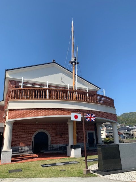 Photo of outside of museum. It is made of red brick and shaped a bit like a boat, with a deck on the upper floor. It has a mast. A flag pole in front of the building has the Japanese and British flags