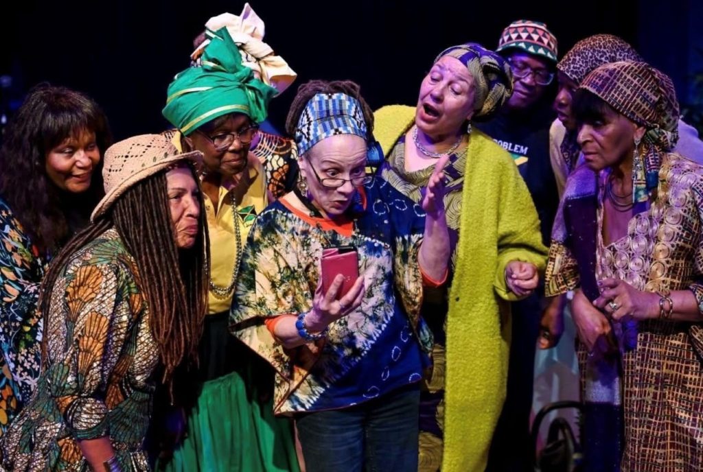 A group of people crowded around one lady who is wearing glasses and hold a phone.  The people are all wearing colourful clothes, headscarves and hats.  