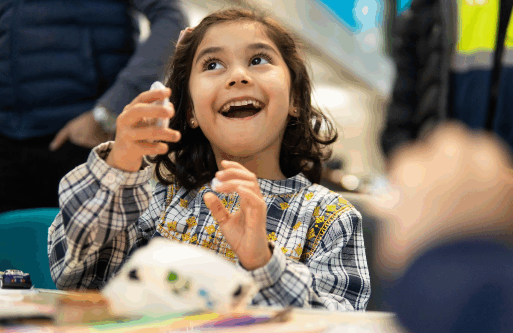 A little girl looking up with her mouth open.  She is wearing a grey  checked top and is sat at a table.