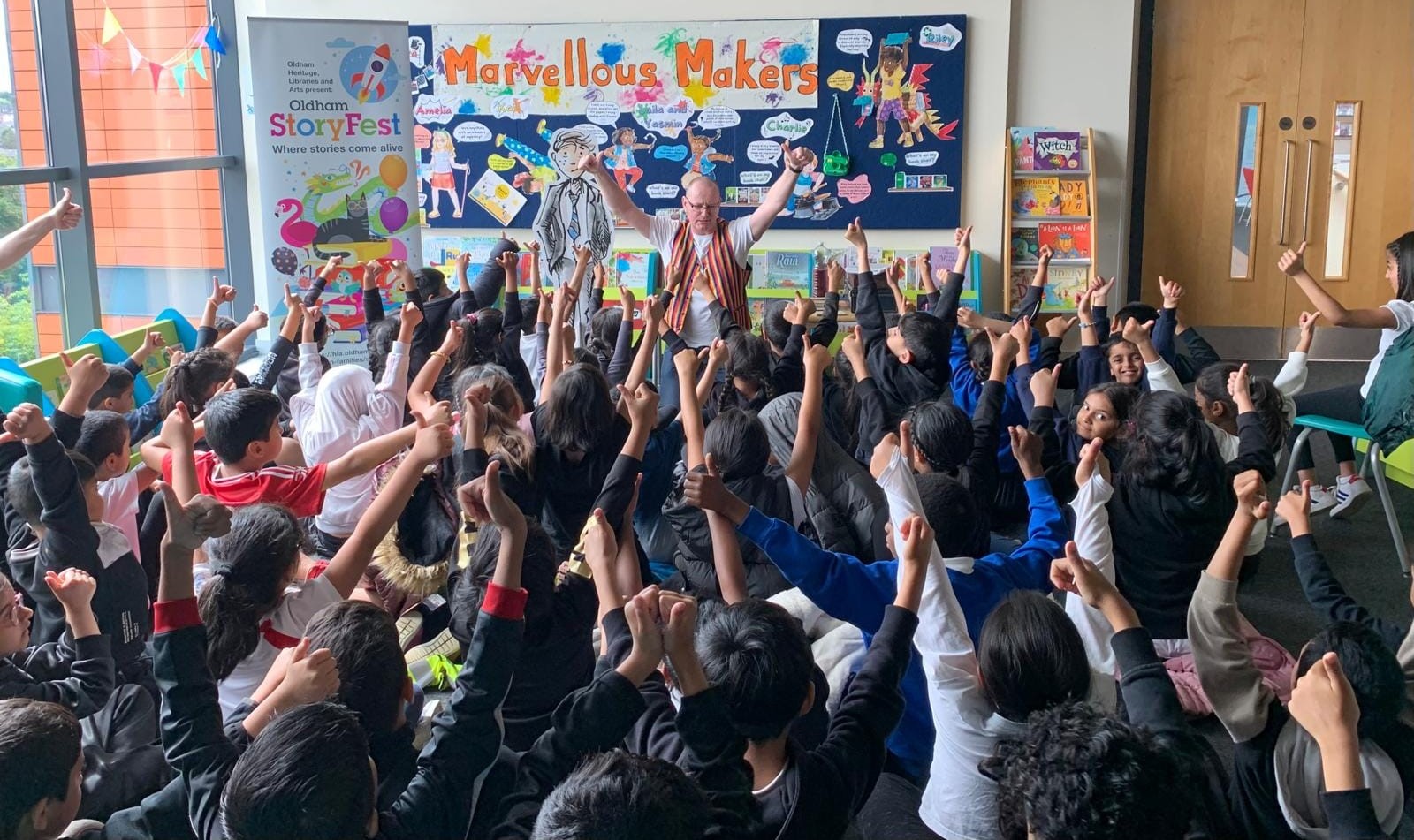 A class of school children facing away from the camera with their arms in the air and their thumbs up.   A man, wearing a stripey waistcoat facing the camera, looking at the children.