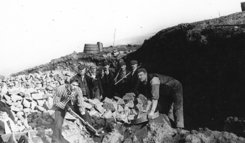 A black and white photograph of a historic dig at Castleshaw Roman Fort, with 7 smartly dressed men shoveling rocks