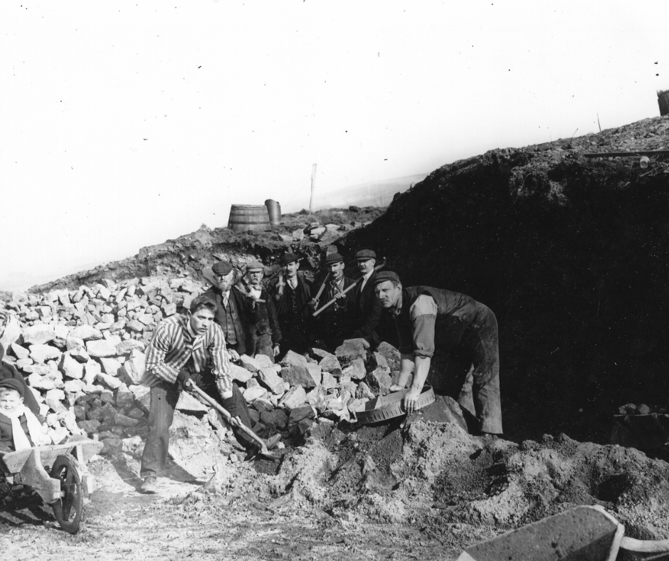A black and white photograph of a historic dig at Castleshaw Roman Fort, with 7 smartly dressed men shoveling rocks