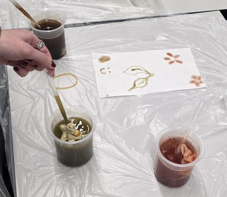 A hand holding a paintbrush in a pot of green dye next to a pot of red and black dye. Flowers, leaves and a smiley face have been painted on a piece of paper with the dyes.