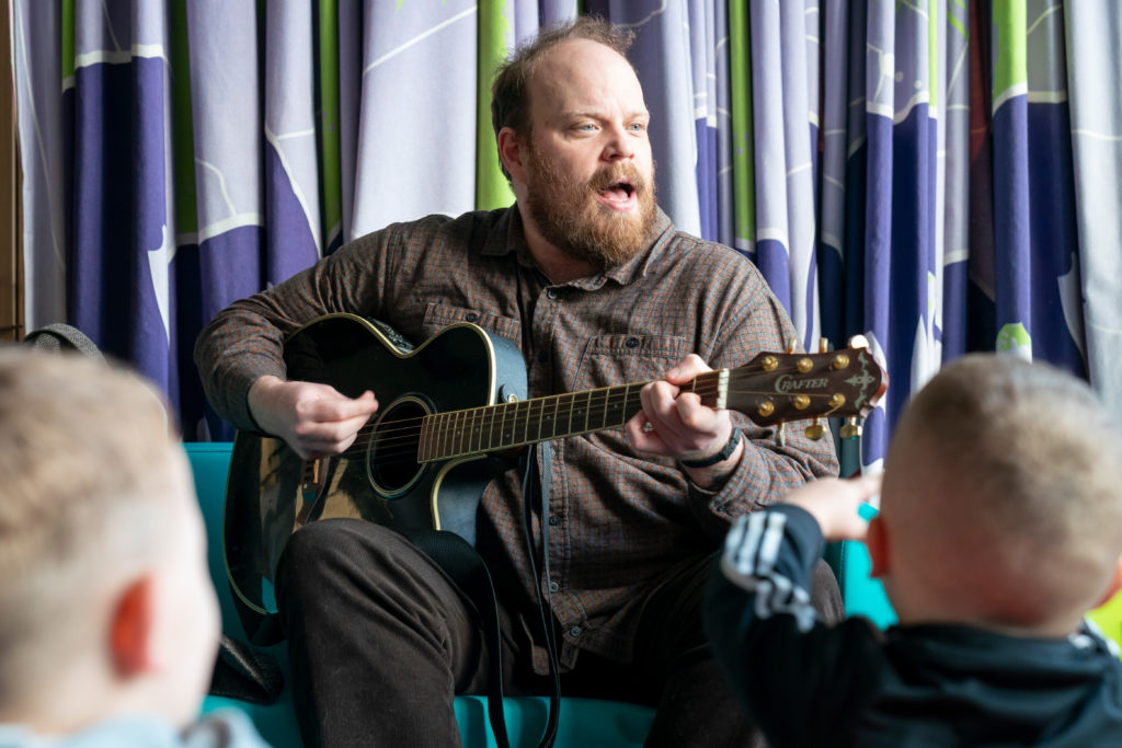 A man with a beard, singing and playing a guitar