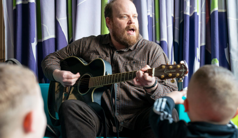 A man with a beard, sat down, singing and playing a guitar