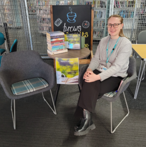 A smiling woman sits at a round table. There's a pile of books on the table and an empty chair to the other side of the woman - the reading for wellbeing drop-in.