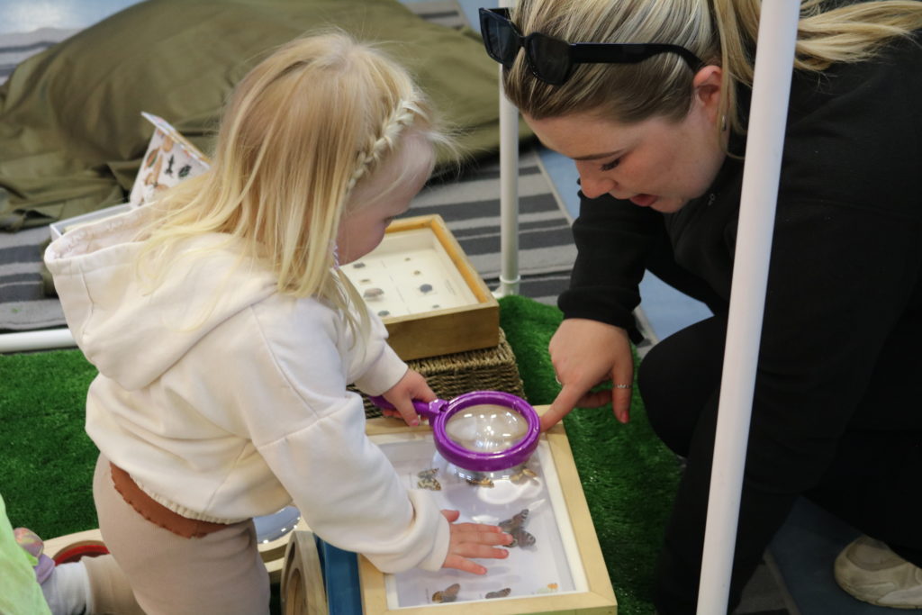 A little girl in a white hoody with a magnifying glass looking at some butterflies with a lady dressed in black