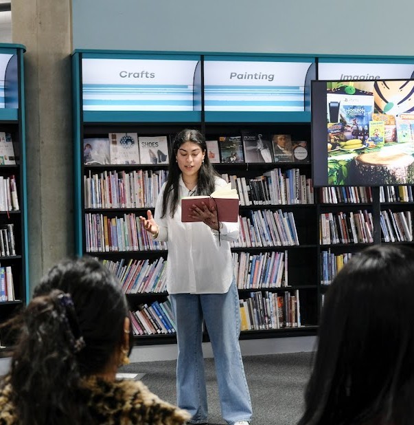 A lady stood near some book shelves speaking to an audience