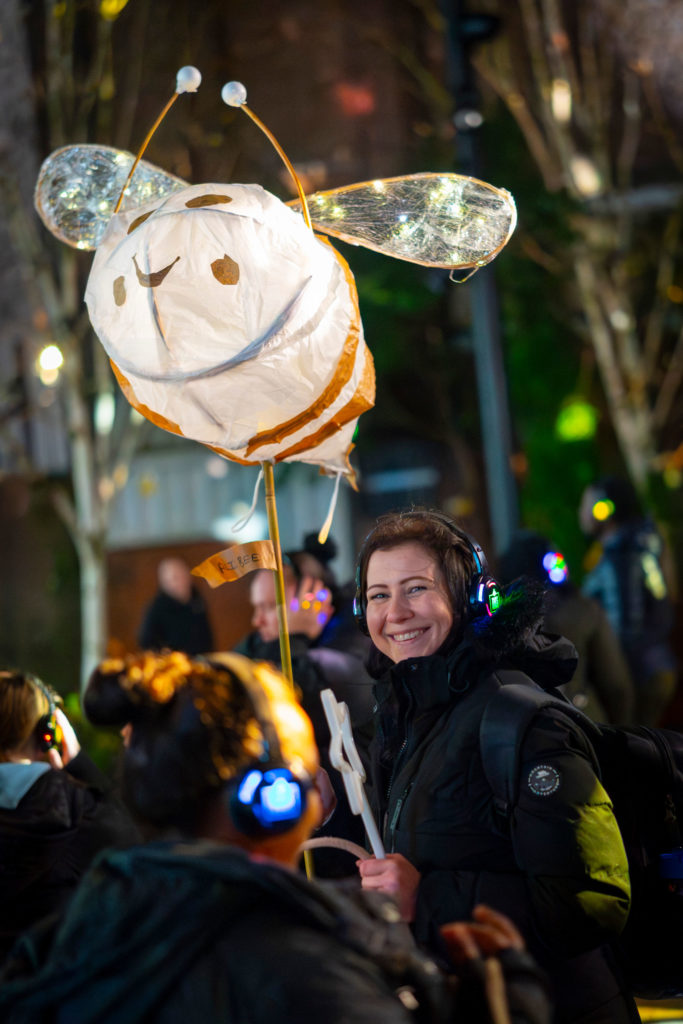 A girl with heaphones holding an illuminated bee on a stick