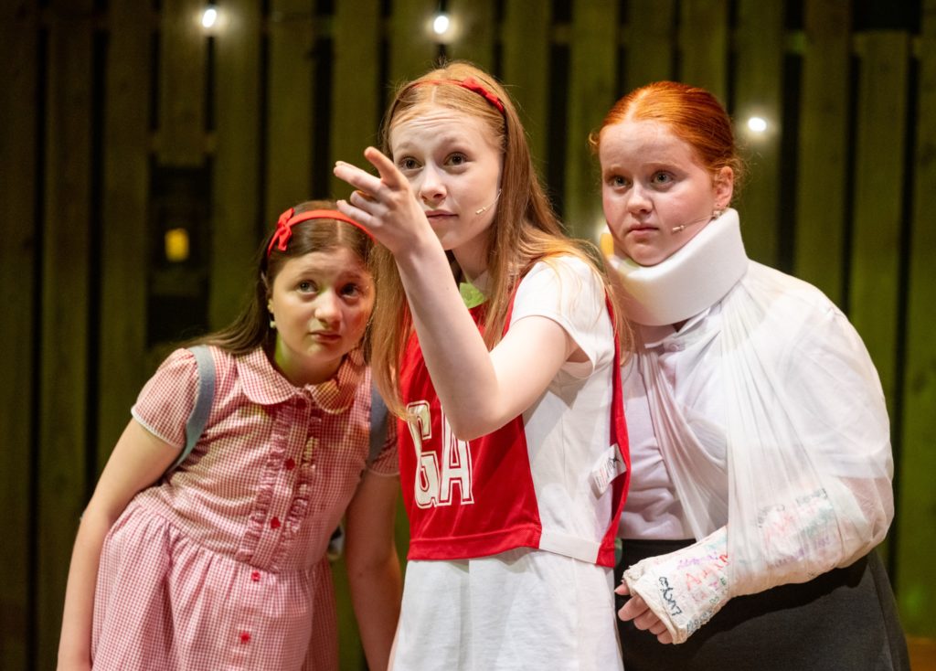 3 young girls looking up. The left hand girl is wearing a headband and red gingham dress, the middle girl is pointing upwards and the girl on the right has her arm in plaster and neck in a brace.