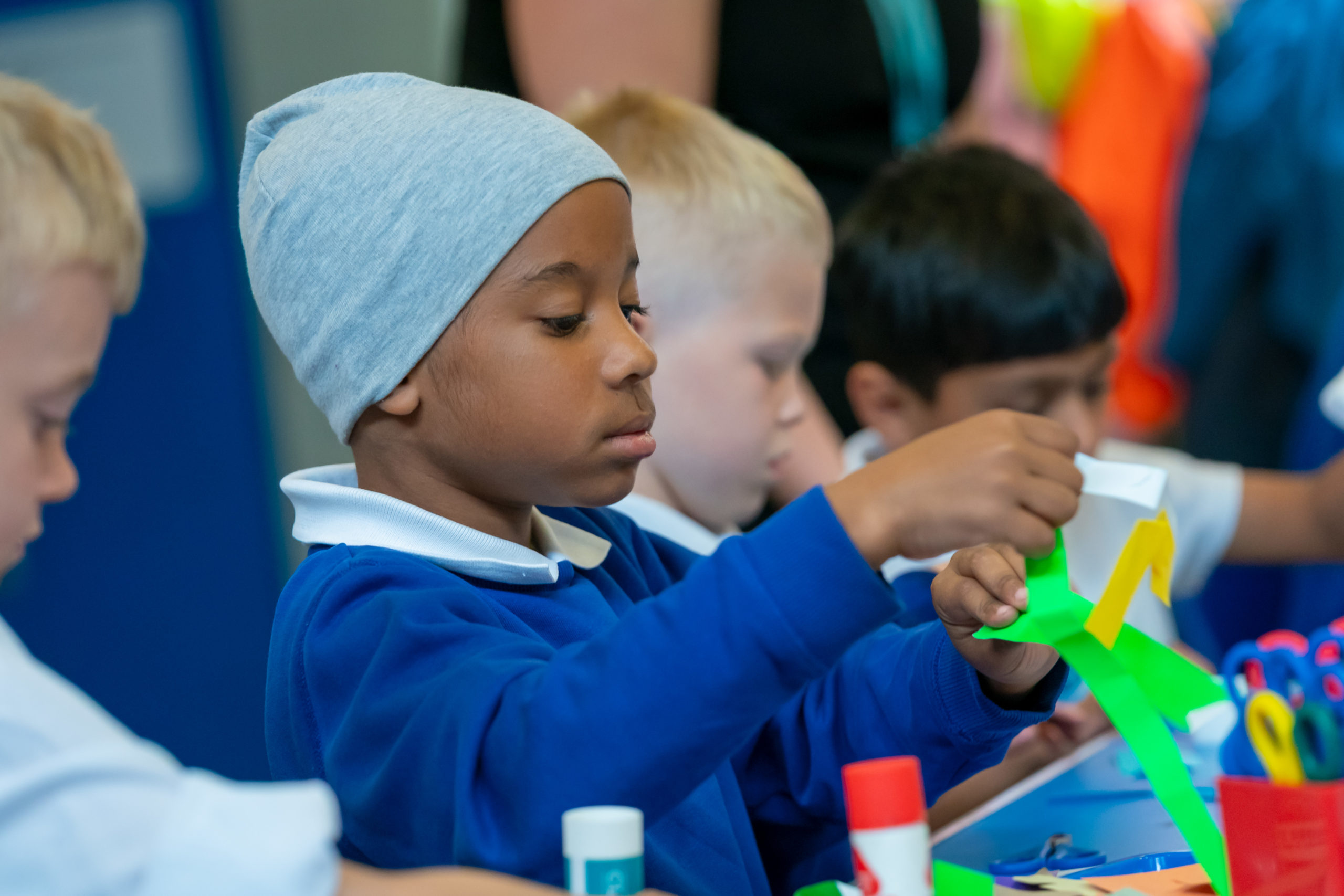 A little boy in a school uniform with a grey beany hat on, making some craft