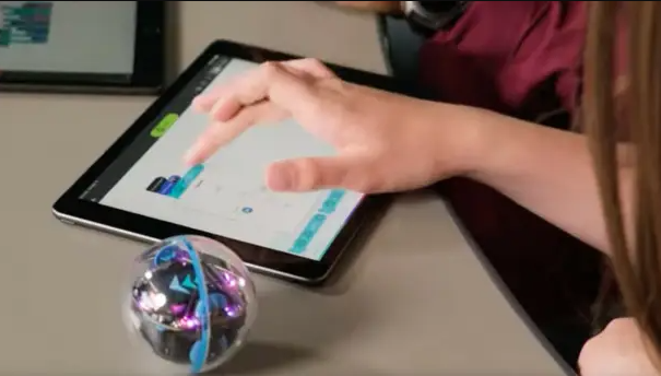 Stock image of a child with long hair (mostly off screen) using a tablet with a Sphero (small ball robot) nearby