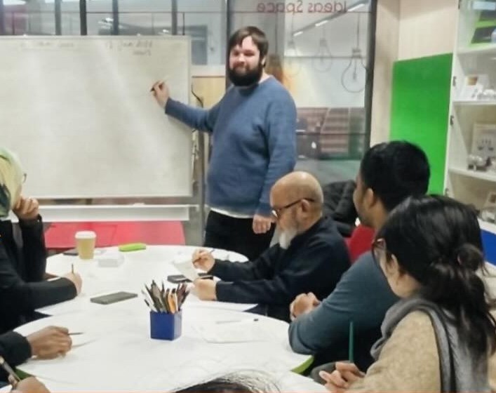A man with a beard writing on a white board. He is looking at some people sat at tables who are writing.