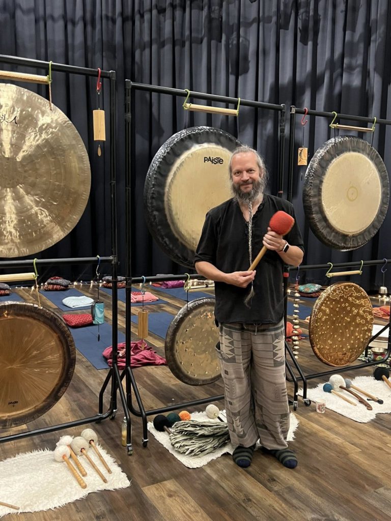 A man with a beard holding a drum stick stood in front of some gongs.