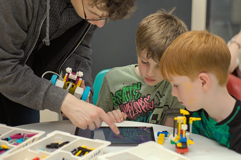 A man showing 2 boys something on a tablet which is on a table with some lego.
